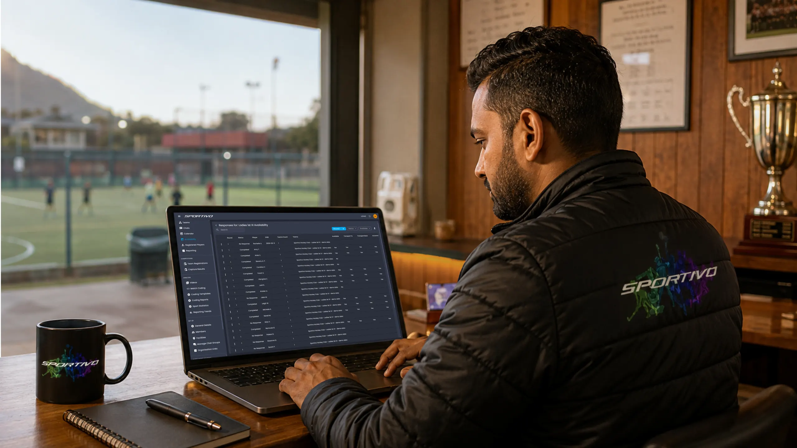 Club manager reviewing team availability responses on a laptop in a clubhouse overlooking the pitch