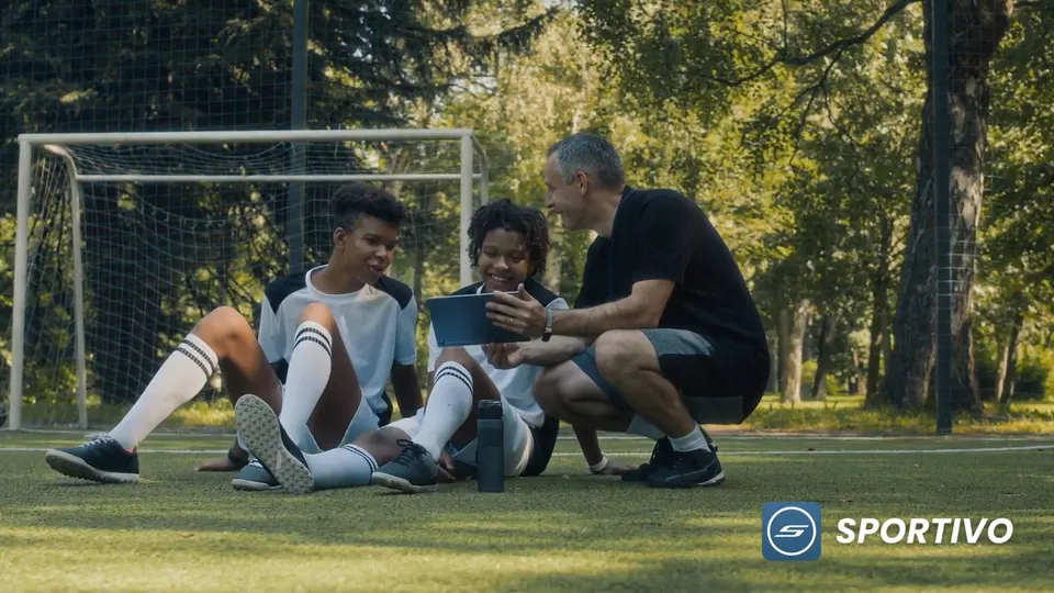 A coach crouches beside two young footballers sitting on a pitch, the three of them looking together at a tablet.